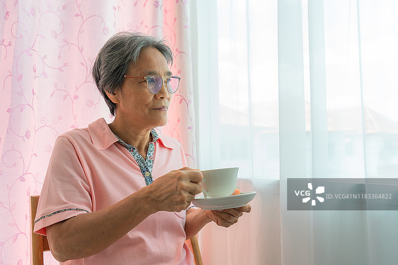 亚洲资深妇女坐在窗户拿着一杯咖啡在客厅在家里图片素材