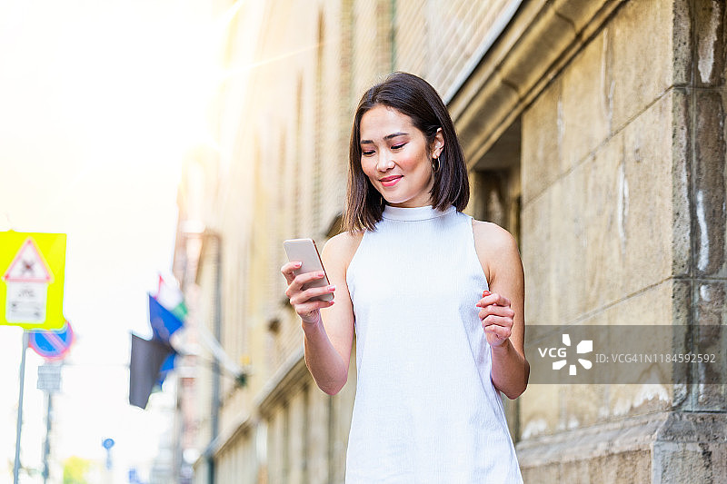 美丽的亚洲女人使用一个应用程序发送短信在她的智能手机设备。快乐的年轻亚洲女人拿着智能手机站在街上图片素材