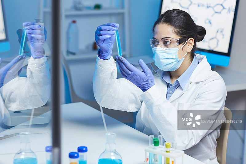严肃的年轻印度女化学家穿着实验服戴着医用口罩坐在桌子前用液体物质做实验图片素材
