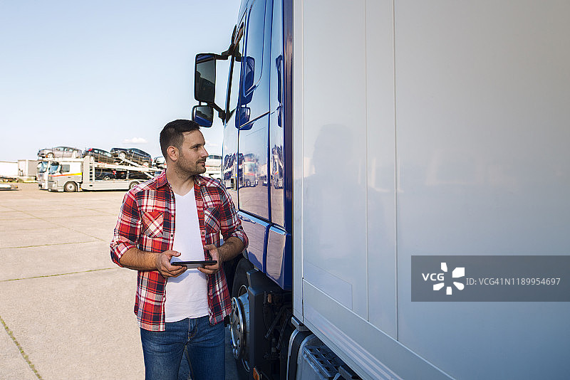 穿着便服的卡车司机站在他的卡车旁，手里拿着写字板，看着油布。运输服务。图片素材