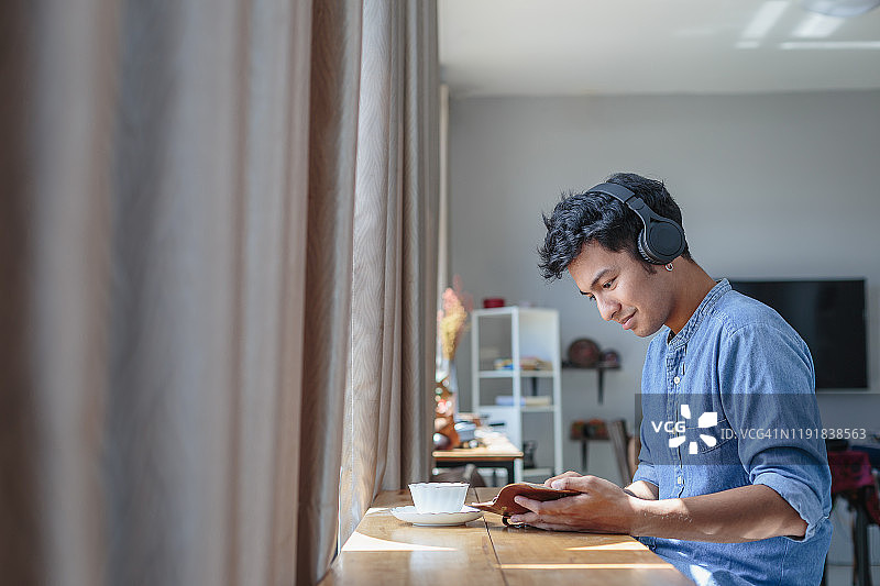 年轻的亚洲帅哥戴着耳机听音乐，在家看书，人们放松的生活方式和科技理念图片素材