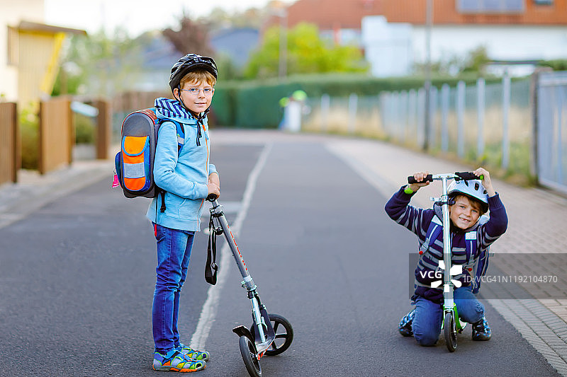 在阳光明媚的日子里，两个学童戴着安全帽，背着背包骑着滑板车在城里兜风。快乐的孩子们穿着五颜六色的衣服骑着自行车去学校。图片素材