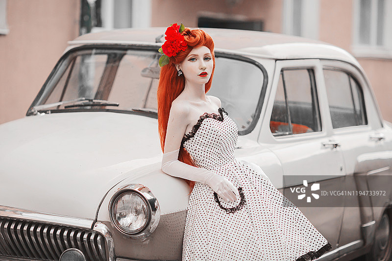 优雅的红发女孩在汽车背景白色复古连衣裙。红唇美女。戴着长手套的女士。城市里戴着手套的时尚复古模特。红头发的女人。复古的车图片素材