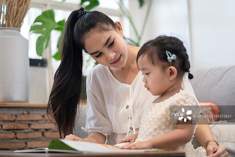 亚洲漂亮的年轻母亲和她的女儿一起读故事书。妈妈高兴地看着小女孩。良好的母子关系。家庭活动与教育理念。图片素材