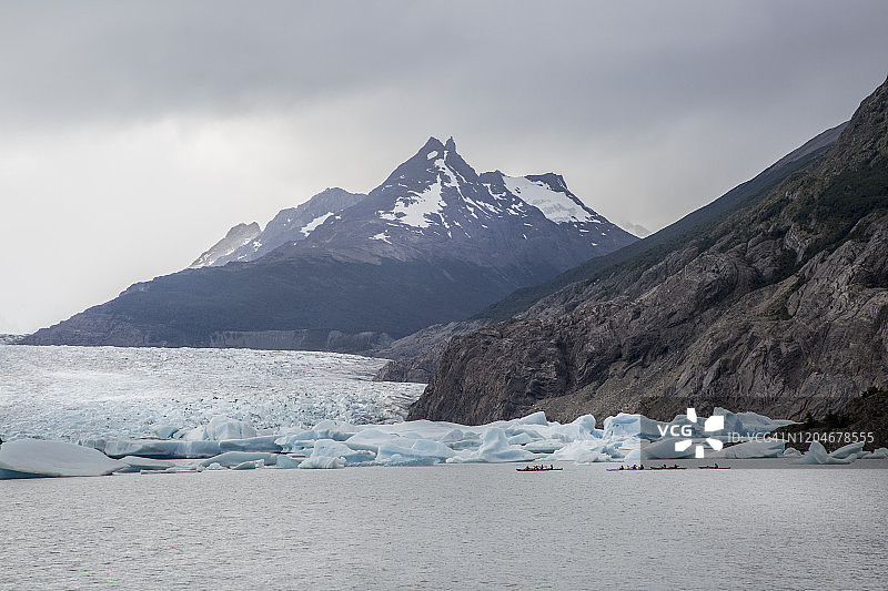 宽阔的镜头，游客划着皮划艇在冰川流入湖泊与积雪覆盖的山脉前。图片素材