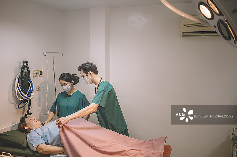 在医院的手术室里，一名亚裔华裔男医生、外科医生和女护士正在为手术做准备，并用毯子盖住病人图片素材