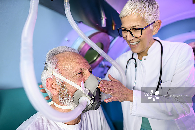 一位灰白头发的女性医生安抚病人，当他坐在高压氧室在HBOT诊所呼吸面罩图片素材