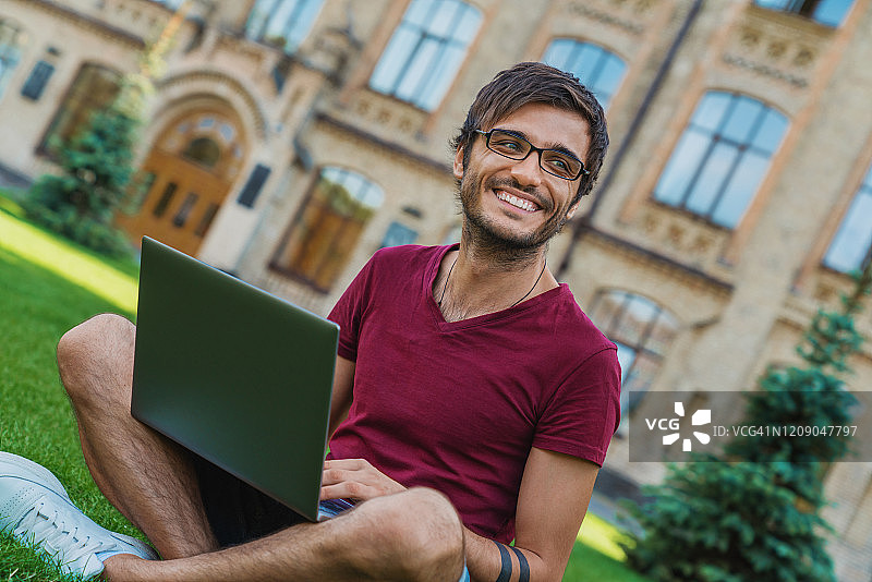 年轻的混血儿男学生戴着眼镜和笔记本电脑坐在大学附近的草地上图片素材