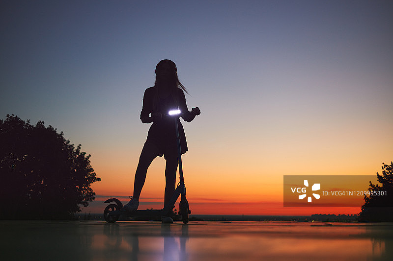 黎明时分，年轻女子骑着电动踏板车的剪影。图片素材