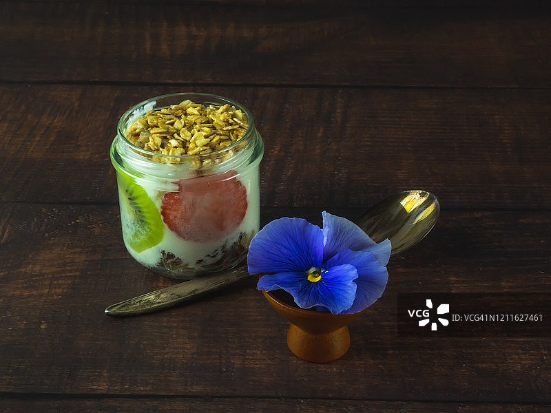 一杯含有猕猴桃、草莓和格兰诺拉麦片早餐的天然酸奶，一朵可食用的花，一把旧羊驼或银勺子放在木桌上图片素材
