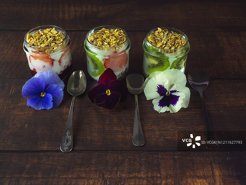 木桌上放着一杯天然酸奶、草莓、猕猴桃、格兰诺拉麦片早餐、可食用的鲜花和旧银勺图片素材