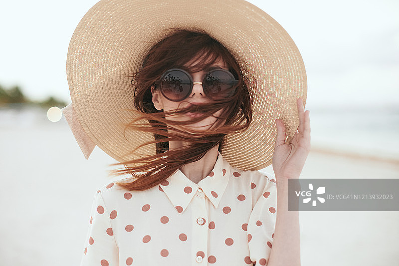 一名年轻女子扔下一件米兰连衣裙和一顶大帽子，头上戴着同样的太阳镜，手里拿着一顶帽子，她的头发被风吹散了。图片素材