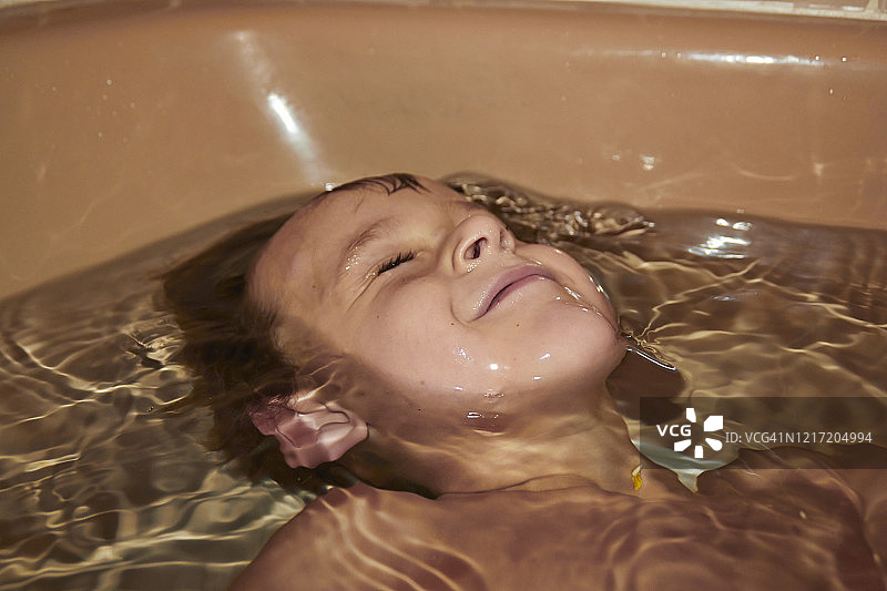 小男孩棕色卷发棕色眼睛在浴缸与半头在水桃子色浴缸赤膊洗澡闭上眼睛进入水图片素材