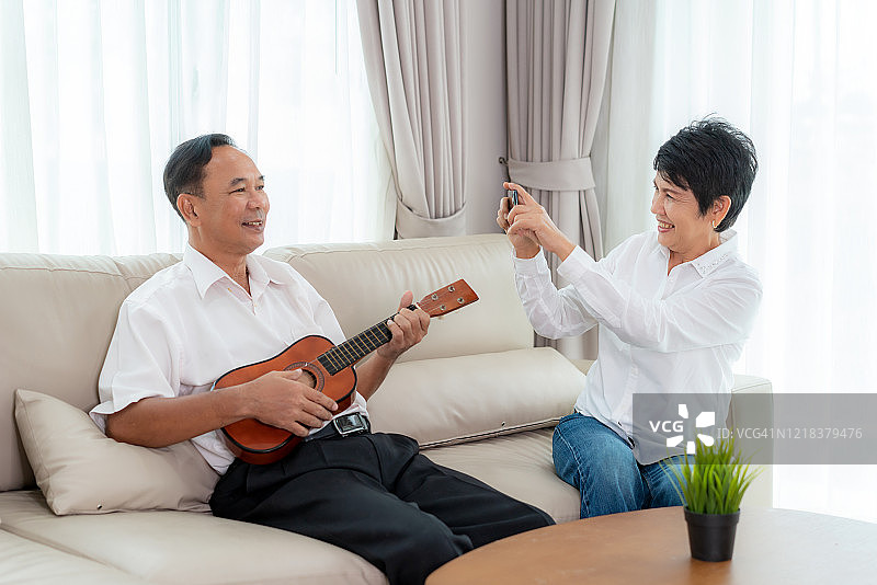 快乐的亚洲老人坐在客厅的沙发上弹奏着尤克里里琴唱歌，而他的老妻子在家里用智能手机录制视频或在社交媒体上直播。图片素材