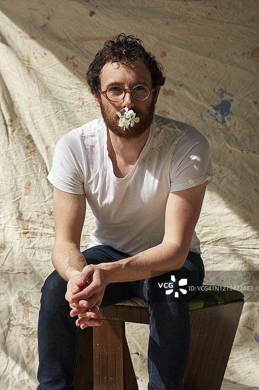 一个男人坐在外面，靠在一块涂了颜料的布上，嘴里叼着一朵花，对着相机看。图片素材