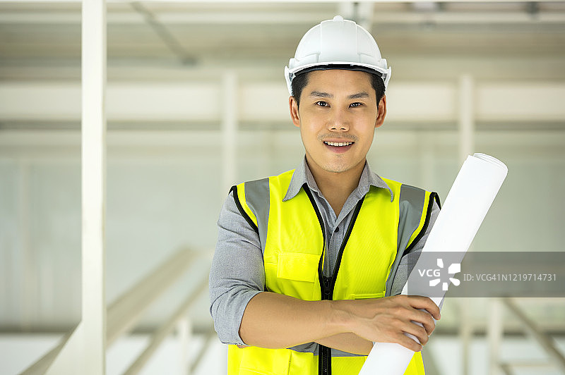 成熟的亚洲工程师穿着安全防护服，手里拿着设计图站在建筑工地前。建设,建筑活动。图片素材