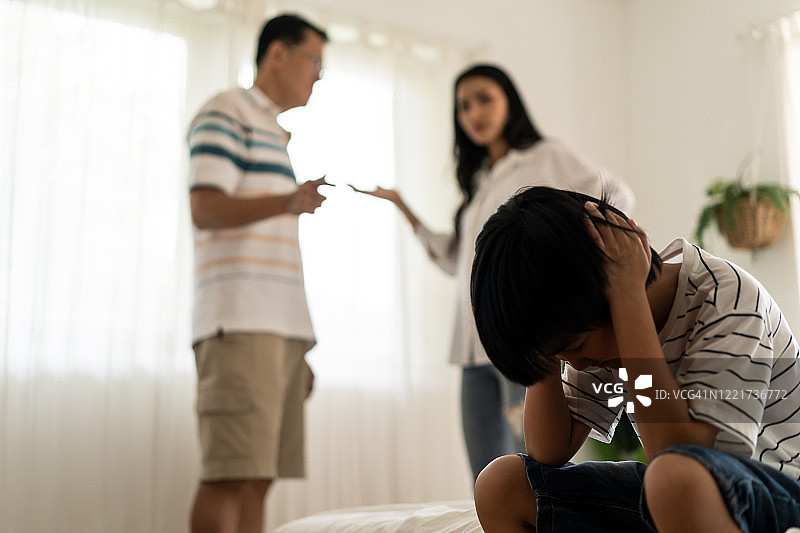 亚洲男孩坐在床上哭泣，而父母在家里打架或争吵冲突。孩子用手捂着耳朵不想听到暴力的争论。家庭中的家庭问题。图片素材