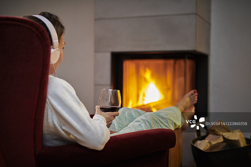 一个女人坐在扶手椅上，拿着一杯红酒，戴着耳机在壁炉前听音乐图片素材