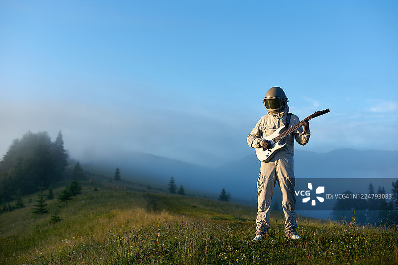 清晨，穿着太空服的宇航员拿着吉他站在美丽的青山草地上图片素材