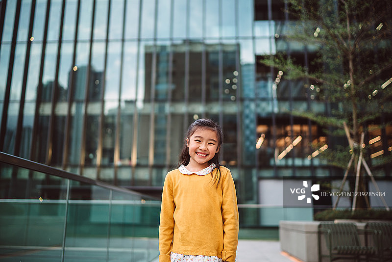 一个可爱的小女孩对着镜头快乐的微笑，在城市的现代建筑图片素材