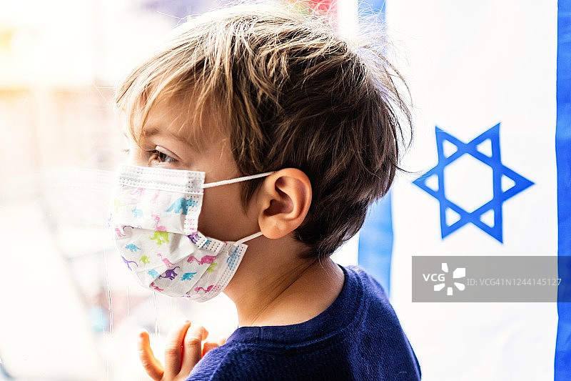 一个小男孩戴着医用口罩从一扇窗户往外看背景是一面以色列国旗图片素材