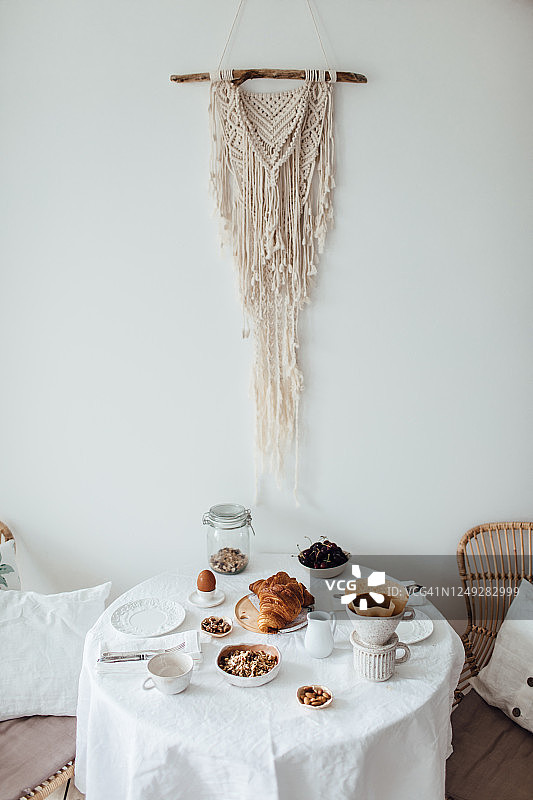 舒适的早餐在家里提供。早晨优雅的餐桌与白色亚麻桌布，并提供健康的早餐在上面。舒适和时尚的室内与棉流苏装饰的背景。图片素材
