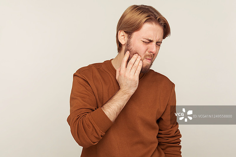 牙齿的健康问题。一幅长胡子的男人的肖像，穿着运动衫，捂着酸痛的脸颊，因剧烈的牙齿疼痛而皱着眉头图片素材