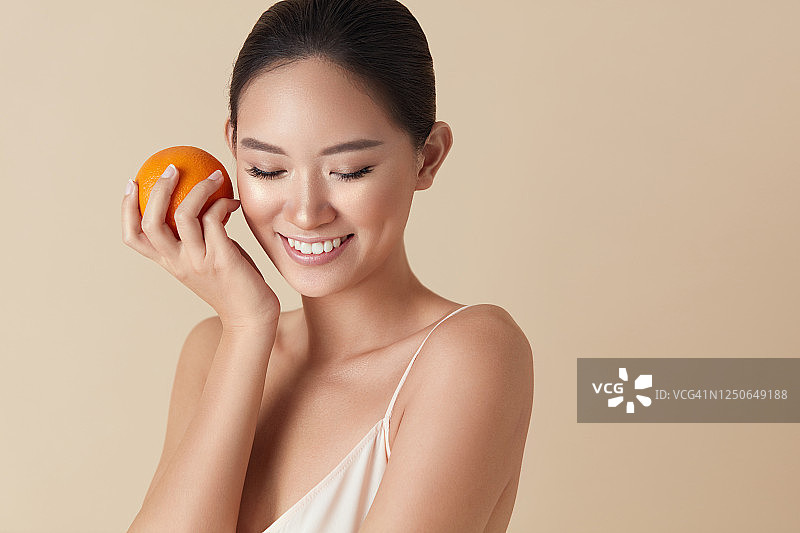 美丽的脸。水果和模特肖像。微笑的亚洲女人手里拿着多汁的橘子，向下看。健康皮肤的天然维生素。美丽的民族女孩与柑橘在米色背景。图片素材