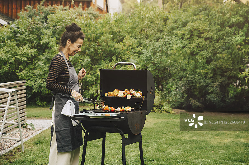 在花园聚会期间，微笑的女人在后院烧烤做饭的侧面视图图片素材