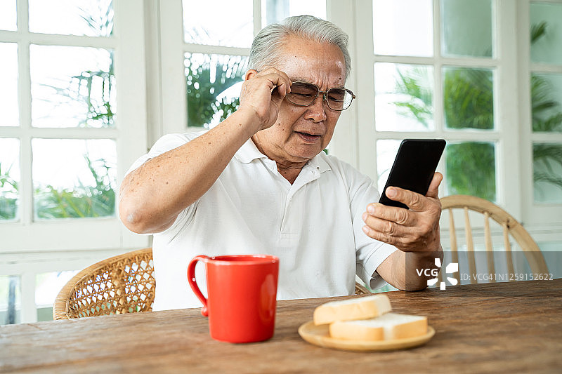 年长的亚洲男性有视力问题，在家里吃早餐时戴着眼镜盯着智能手机看信息。在室内视力模糊的老人图片素材