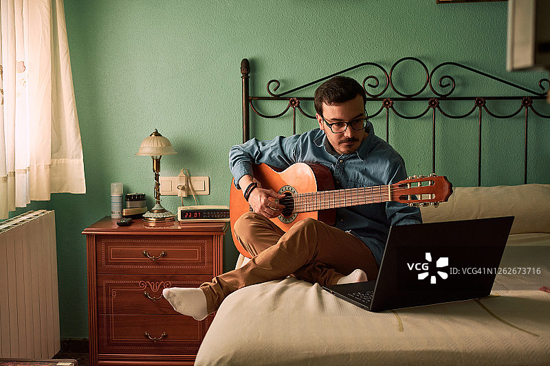 一个年轻人在家里通过看笔记本电脑练习弹吉他图片素材