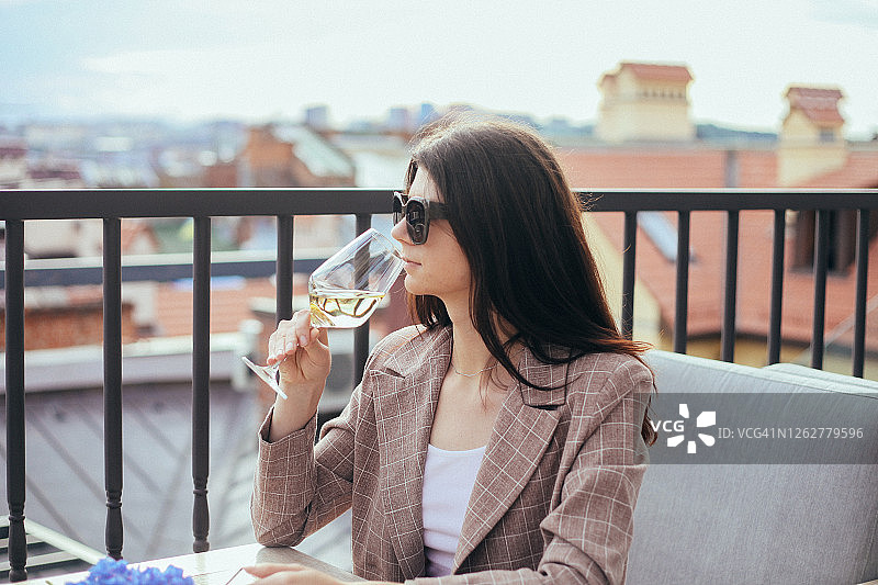 一个女人坐在餐厅屋顶上喝着白葡萄酒，拿着酒杯。夏季旅游、休闲、商务和活动概念。图片素材