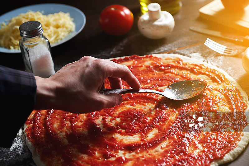 在家里的木桌上用蔬菜和蘑菇烹饪意大利素食披萨。一个女人用勺子在面团上涂番茄酱。一步一步的指导，自己做。步骤3。图片素材