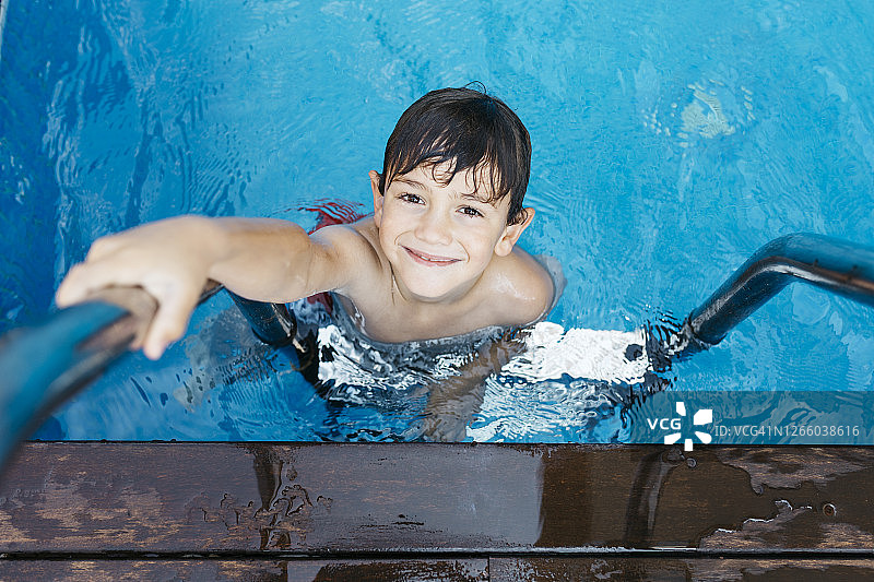 微笑的男孩拿着梯子站在游泳池里图片素材