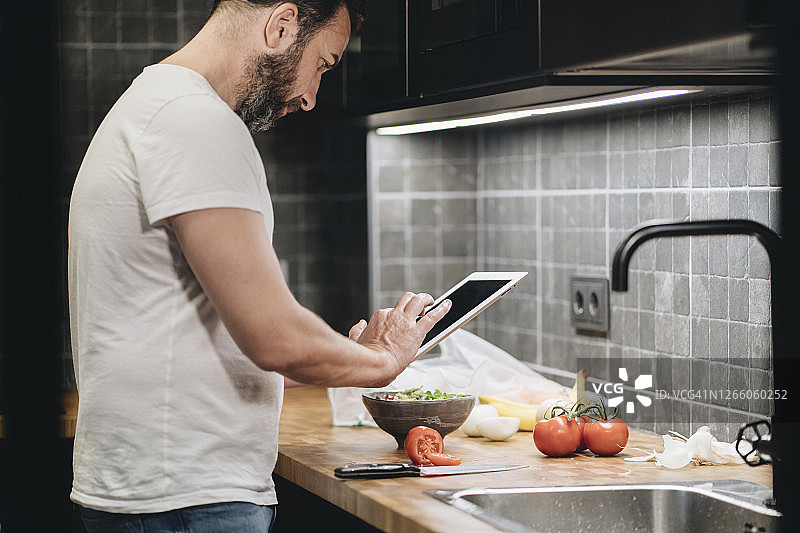 成熟的男人站在厨房里，准备食物，使用网上菜谱图片素材