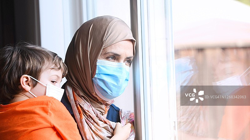 一名成年穆斯林妇女戴着头巾和防护面罩，抱着儿子从窗户往外看图片素材