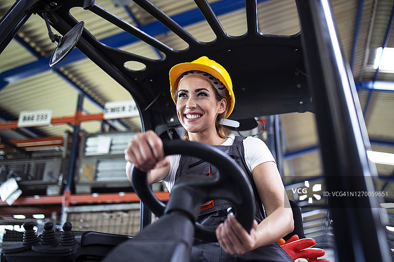 美丽微笑的女工人在工厂仓库驾驶叉车。图片素材