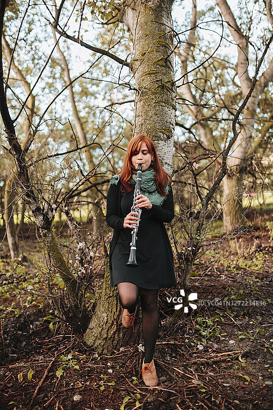 美丽的年轻女子练习单簧管时，倚在光秃秃的树干在森林图片素材