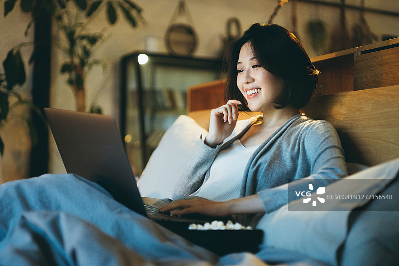 一名年轻的亚洲女子躺在床上享受周末，在笔记本电脑上看电影，晚上在家吃爆米花图片素材
