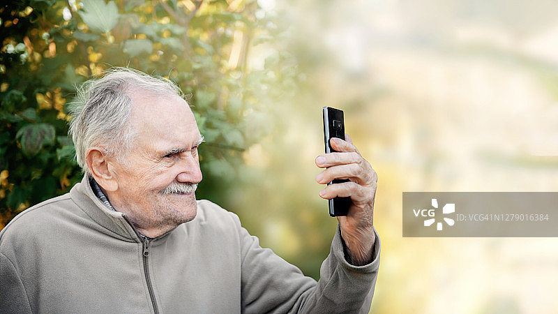 上了年纪的活跃的英俊男子用一部手机自拍。一位老人看着镜头，一边说一边笑。复制空间,旗帜。图片素材