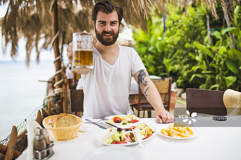 一个留着胡子的年轻人在海边的餐馆里喝啤酒图片素材