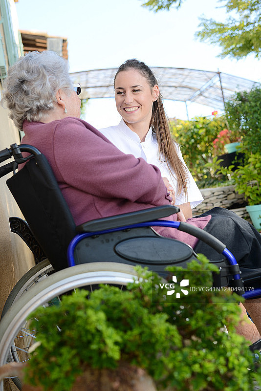 老年妇女在轮椅上与护士在户外护理之家医院花园。图片素材
