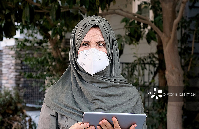 成熟的白人穆斯林妇女拿着她的数字平板电脑，戴着n95保护面罩图片素材