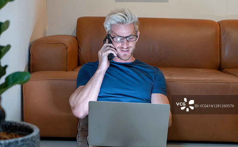 年轻迷人的微笑的家伙正在浏览他的笔记本电脑和聊天哦手机，坐在家里舒适的沙发上，穿着休闲装。自由职业的商业工作在家的概念图片素材