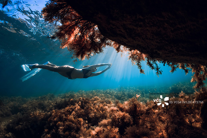 有白色鳍的自由潜水女孩在阳光下的岩洞附近滑行。在水下自由潜水图片素材