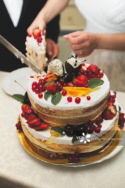 新娘和新郎在白色的木桌上切了一个优雅的用浆果装饰的两层婚礼蛋糕。图片素材