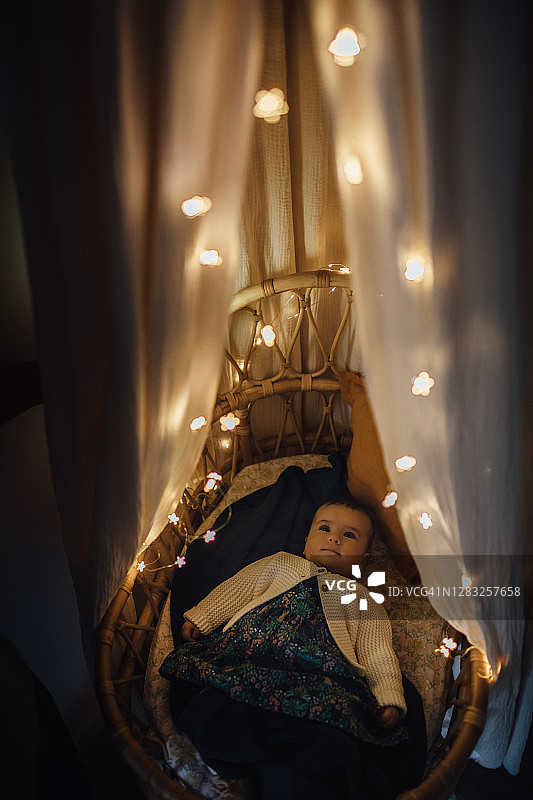 睡意朦胧的男孩在舒适的婴儿床里打盹，家里有顶棚和花环。高角度的婴儿睡觉与照明灯的藤条摩西篮子。圣诞装饰。图片素材