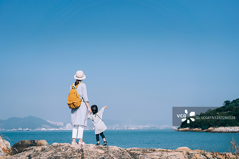 背影是年轻的亚洲母亲牵着女儿的手爬山，背靠蓝天和大海。他们玩得很开心，一起度过了一段高质量的亲密时光。在一个阳光明媚的日子里，享受大自然和美丽的风景图片素材