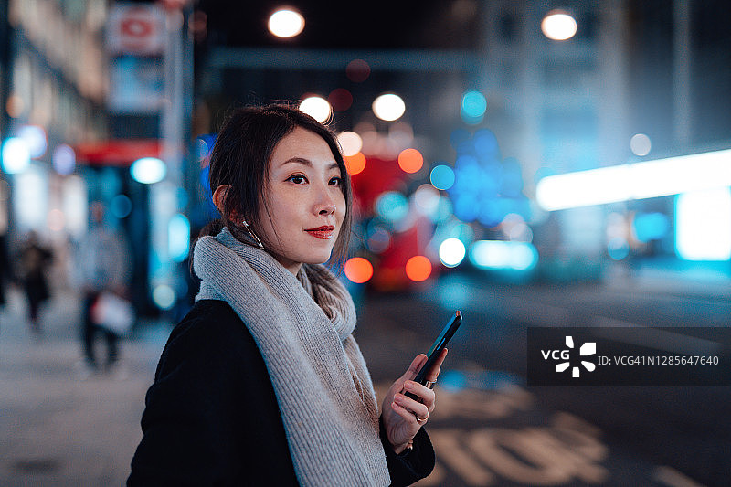 年轻女子用智能手机在晚上的城市里等出租车图片素材