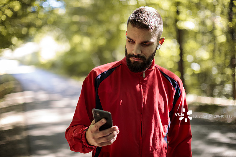 年轻的运动员肖像与蓝牙耳机使用手机，而在森林慢跑图片素材
